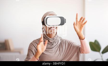 Black lady in traditional hijab using VR headset to interact with virtual reality at home Stock Photo