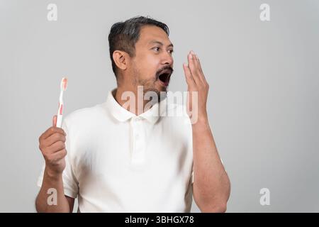 Middle-aged Asian man checking his breath with hand near mouth while holding toothbrush, concept of oral hygiene, bad breath problem, morning routine, Stock Photo