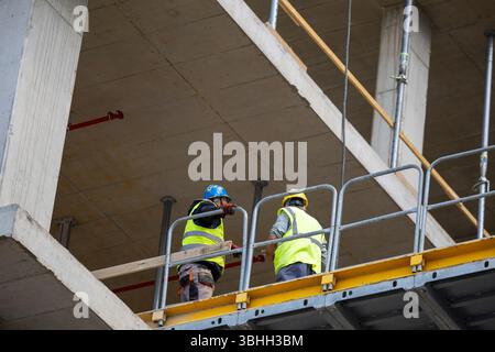 Two male workers engage in discussion while standing on scaffolding at construction site, focusing on their tasks under clear sky. Stock Photo