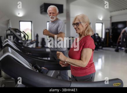 Smiling older woman and man doing their daily cardio, jogging on treadmill Stock Photo