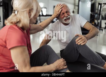 Worried senior woman petting husbands head in comfort after knee injury during workout in gym Stock Photo