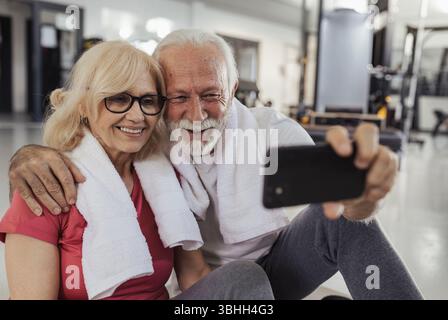 Satisfied senior couple resting on gym floor, taking pictures with smiles on their faces Stock Photo