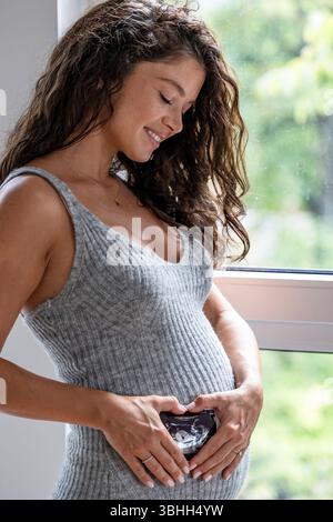 Smiling pregnant woman with ultrasound image in front of her stomach. Her hands forming heart shape Stock Photo