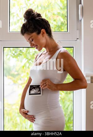 Pretty pregnant woman posing in front of a window as she holds both the ultrasound image and her big belly Stock Photo