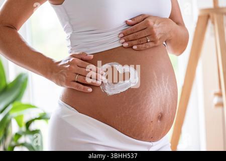 Close up of woman drawing a heart with sunscreen on her pregnant belly, showing love for her baby Stock Photo
