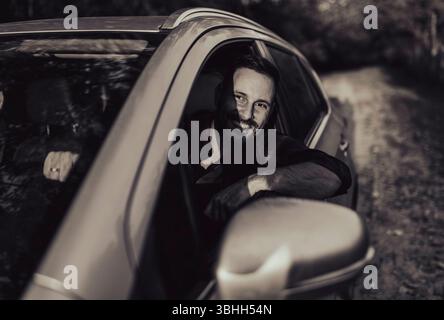 Man driving car, smiling while gripping the steering wheel and looking through side window. Bright trees and a sunny day create a cheerful atmosphere Stock Photo