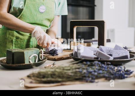 Person stamps lavender soap near dried flowers and green bars in a small workshop. Aromatherapeutic soap, functional wellness, lavender benefits Stock Photo