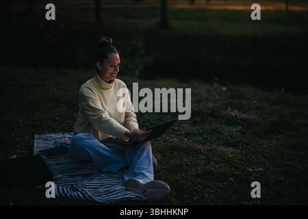 Woman Sitting Outdoors on Blanket Using Laptop During Evening Stock Photo