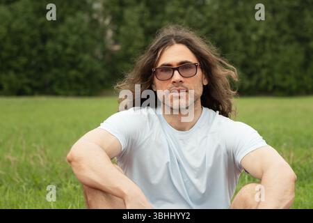 Long-haired man in sunglasses sits and relax on running track, wearing t-shirt and shorts, looking relaxed and thoughtful. Summer outdoor workout. Stock Photo