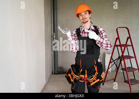 young man builder holding palm open for new product point finger at arm with copy space at construction site Stock Photo