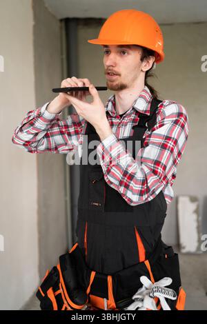 Builder in yellow helmet recording voice message dictating something to online assistant using smartphone and modern technologies Stock Photo