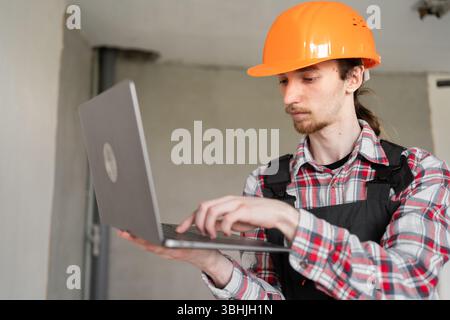 Portrait builder in helmet using laptop at construction site. Young foreman using computer standing in renovating apartment Stock Photo