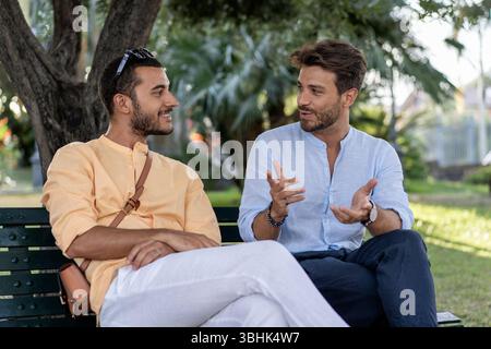Two young male friends or business colleagues having an engaging and positive conversation while sitting on a bench in a city park. A concept of commu Stock Photo