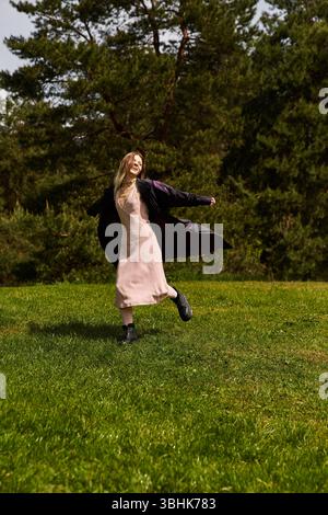 A fair haired woman, wearing a dress and jacket, twirls joyfully in a grassy field on a sunny day. Stock Photo
