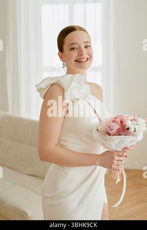 Portrait of young adult Caucasian woman smiling while holding wedding bouquet, standing in bright room during wedding celebration, hair tied back, looking at camera Stock Photo