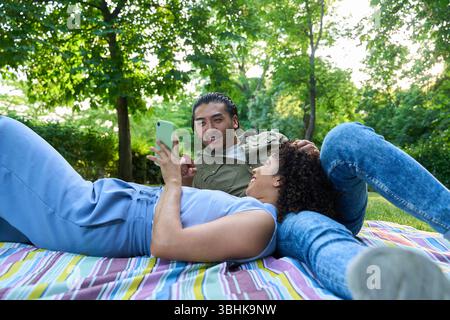 Young multi ethnic couple lying on blanket in park, using smartphone and enjoying leisure time together Stock Photo