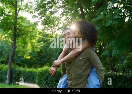 Young multi ethnic couple having fun piggybacking in a green park, enjoying a sunny summer day together Stock Photo