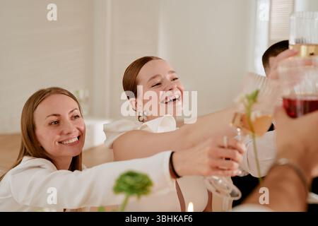 Caucasian young adult women smiling and raising glasses in celebratory toast during wedding celebration, surrounded by friends, joyful expressions visible, hands holding drinks extended Stock Photo