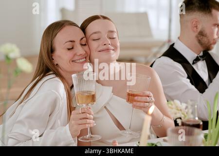 Caucasian young adult bride and Caucasian middle aged woman smiling and embracing while holding wine glasses at wedding celebration, Caucasian young adult man sitting beside them Stock Photo