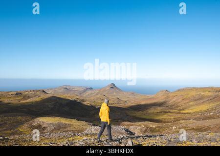 Lone hiker in yellow jacket walks rocky trail under bright blue sky on Snaefellsnes peninsula in Iceland, surrounded by vast volcanic mountains and oc Stock Photo
