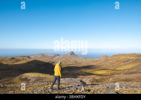 Lone hiker in yellow jacket walks rocky trail under bright blue sky on Snaefellsnes peninsula in Iceland, surrounded by vast volcanic mountains and oc Stock Photo