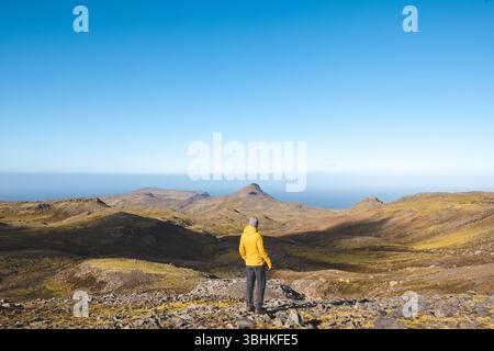 Lone hiker in yellow jacket walks rocky trail under bright blue sky on Snaefellsnes peninsula in Iceland, surrounded by vast volcanic mountains and oc Stock Photo