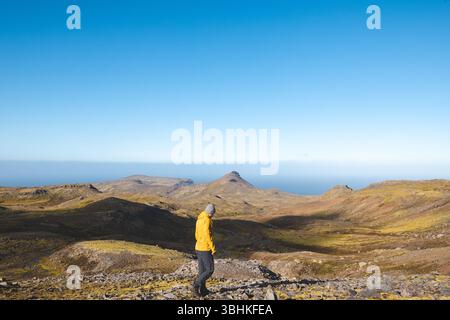 Lone hiker in yellow jacket walks rocky trail under bright blue sky on Snaefellsnes peninsula in Iceland, surrounded by vast volcanic mountains and oc Stock Photo