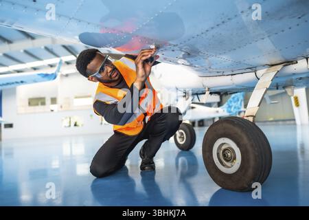 African american aviation technician wearing safety glasses and vest inspecting aircraft wing in hangar Stock Photo
