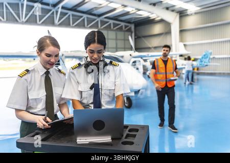 Two female colleagues pilots preparing flight plan using laptop and writing on clipboard in hangar with aircraft and airplane mechanic Stock Photo