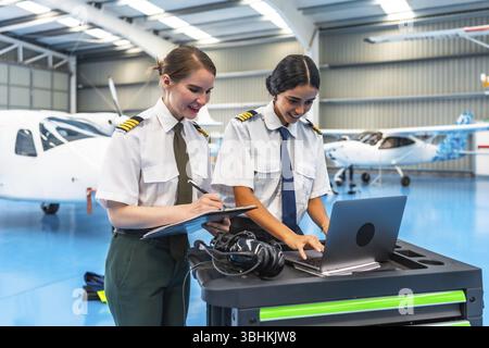 Two female pilots in uniform working on a laptop and clipboard in a spacious aircraft hangar, surrounded by small planes Stock Photo
