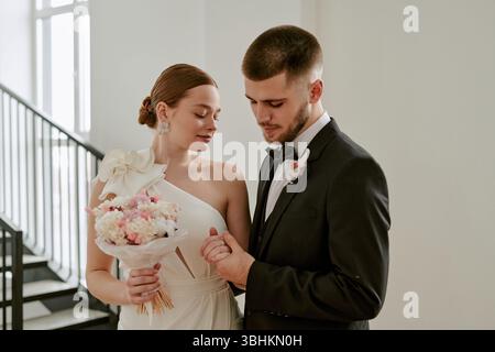 Caucasian young adult woman in wedding dress holding bouquet standing beside Caucasian young adult man in suit holding her hand during wedding celebration indoors Stock Photo