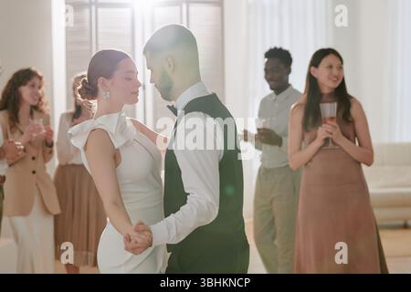 Caucasian young adult bride and groom dancing closely during wedding celebration, multiethnic group of young adult guests standing in background holding drinks and watching couple Stock Photo