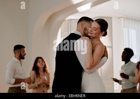 Caucasian young adult bride embracing Caucasian young adult groom while dancing at wedding celebration, multiethnic group of young adults standing in background socializing Stock Photo