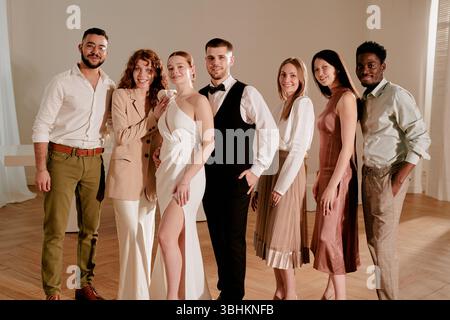 Group of young adult and middle aged multiethnic men and women standing together smiling at wedding celebration, bride and groom posing with friends in elegant indoor setting Stock Photo