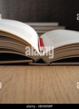 A book is open to a page with a pencil on it. The pencil is red and is leaning on the book. The book is ready for reading or study, with more books bl Stock Photo