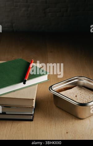 A stack of books and a pencil on a table. The pencil is red and the book on the top is green. Metal lunch box  on the side. Back to school Stock Photo