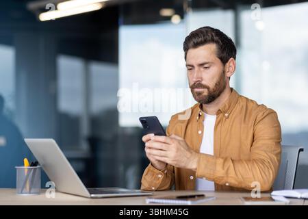 A focused man uses his smartphone while sitting at a desk in an office setting. Laptop and pen holder on the table. Stock Photo