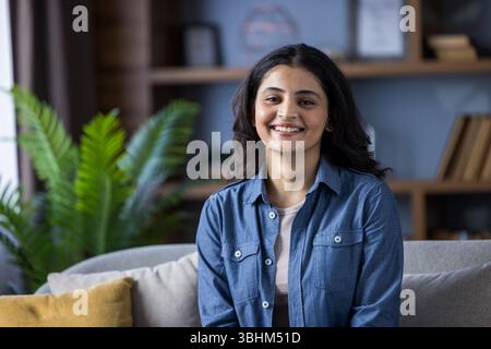 A beautiful young woman sits comfortably at home, smiling radiantly. She is wearing a denim shirt and looks happy and relaxed. Stock Photo