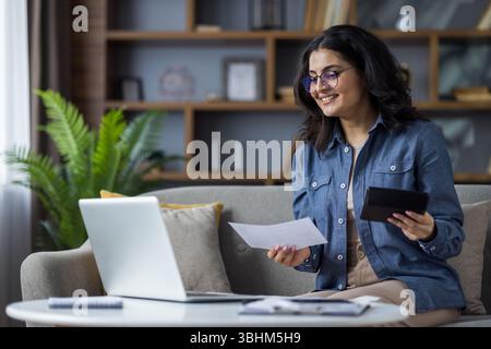 A smiling young woman works from home, holding documents and a tablet while using her laptop on the couch. Stock Photo