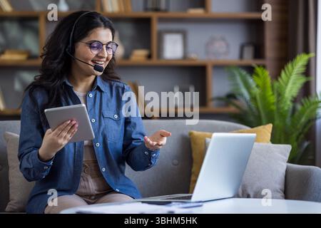 A Muslim woman in a headset sits at home on a sofa in front of a laptop, holds a notepad in her hands and talks via video call to the camera. Stock Photo
