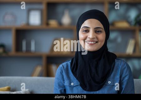 Close-up portrait of a young Muslim woman in a black hijab sitting on a sofa at home and smiling at the camera. Stock Photo