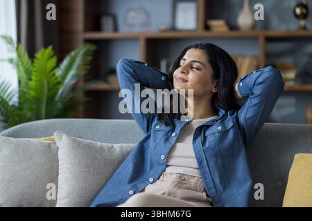 A serene young woman relaxes on a comfortable couch, hands behind her head, and closed eyes. She embodies tranquility in a cozy, home setting. Stock Photo