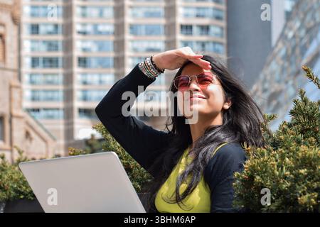 Young woman in city sitting with laptop computer squinting covering eyes from the sun Stock Photo