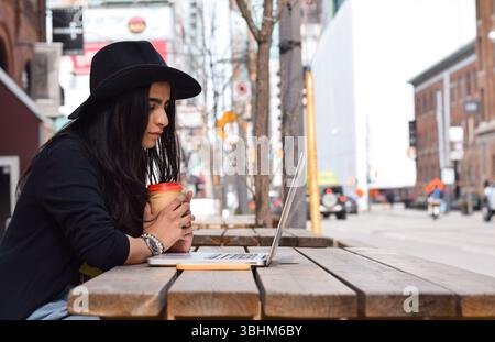 Young woman in fedora hat sitting at outdoor city street cafe using laptop computer Stock Photo