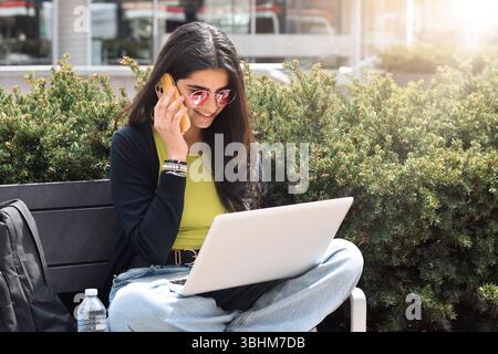 Young woman sitting on street bench using laptop computer and talking on mobile phone Stock Photo