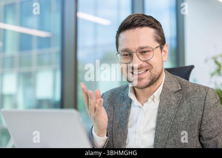 Smiling young male office worker holding online conversation and conference. Sitting in office in front of laptop camera and gesturing with hands. Stock Photo