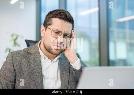 Close-up portrait of a sad and worried young man holding his head and looking at the laptop screen at his workplace in the office. Stock Photo