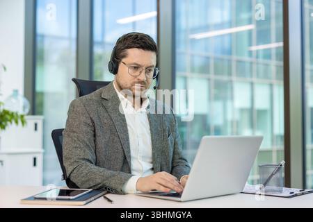 Serious young man in a suit and headset working intently on a laptop while sitting at a desk in the office. Stock Photo