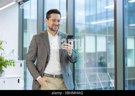 Smiling young male businessman standing in office, holding hand in pocket and using mobile phone. Stock Photo