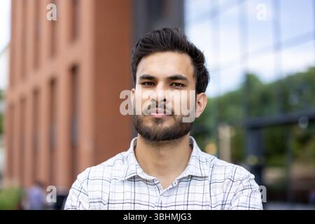 Close-up portrait of a young Muslim man in a shirt standing outside an office building, university campus, looking seriously at the camera. Stock Photo